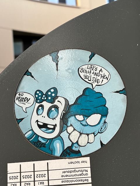 Das Stickerbild zeigt eine runde Illustration mit zwei Comicfiguren auf blauem Hintergrund. Links befindet sich eine Figur mit weißem Gesicht, großen schwarzen Augen, Schleife auf dem Kopf und offenem Mund. Rechts daneben steht eine Figur mit Mütze, rundem Kopf und breitem, gezacktem Grinsen. Beide Figuren haben Sprechblasen mit englischen Texten. Der Rand des Stickers ist leicht beschädigt und zeigt dunkle Risse im Hintergrund.