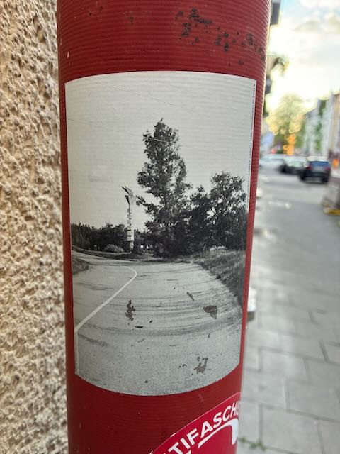 Der Sticker zeigt ein rechteckiges, schwarzweißes Foto. Abgebildet ist eine Straße, die nach links abbiegt. Rechts davon steht ein großer, dicht bewachsener Baum, daneben sind weitere Bäume erkennbar. Am linken Rand der Aufnahme ist ein schmaler Mast oder Pfosten sichtbar. Der Himmel ist hell, der Boden asphaltiert. Das Bild ist ohne erkennbare Personen oder Fahrzeuge aufgenommen. Der Sticker ist auf eine rote Oberfläche geklebt und weist leichte Kratzer und Abnutzungsspuren auf.