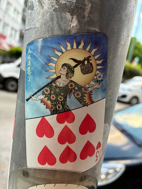 Der Sticker zeigt ein Kartenmotiv, das eine Figur in farbigem Gewand darstellt. Die Person trägt ein Musterkleid, hält einen Stock mit Beutel über der Schulter und eine Blume in der Hand. Im Hintergrund ist eine große gelbe Sonne mit Strahlen zu sehen. Darunter befindet sich ein Kartenmotiv mit roten Herzen in zwei Reihen, das die Zahl 13 symbolisiert. Der Sticker hat abgerundete Ecken und ist rechteckig geformt.