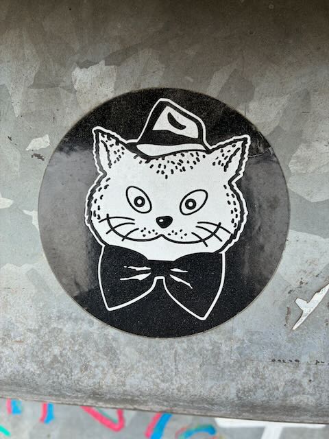 Schwarz-weißer Aufkleber auf grauer Betonwand zeigt eine Katze mit gepunkteten Ohren, großen Augen, Schnurrhaaren, einem Hut auf dem Kopf und einer schwarzen Fliege am Hals, umgeben von einem schwarzen Kreis.