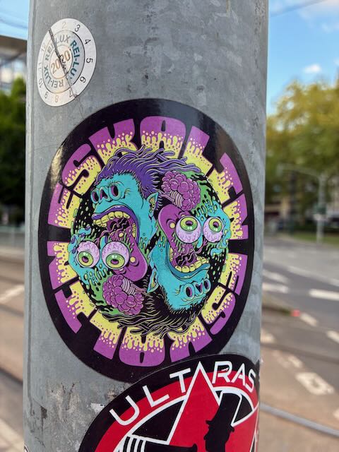 Ein runder Sticker mit einem bunten, comicartigen Motiv zeigt zwei grün-lila Monsterköpfe mit großen, hervorstehenden Augen und offenen Mündern. Um das Bild verläuft der Schriftzug "ESKALATION" in gelb und lila auf schwarzem Hintergrund.