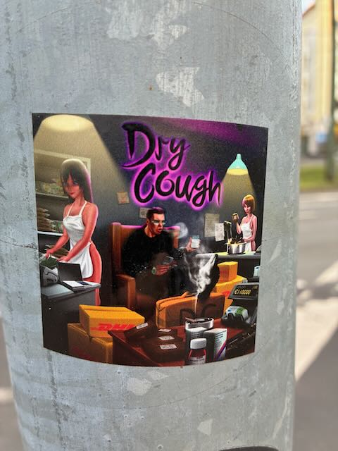Aufkleber mit dem Aufdruck "Dry Cough" im Stil einer Computersimulation: Drei Personen in einem modern eingerichteten Raum, umgeben von Paketen und Technik, eine Figur sitzt auf einem Sofa, andere stehen an Tischen. Auf einem Paket ist das DHL-Logo zu erkennen. 