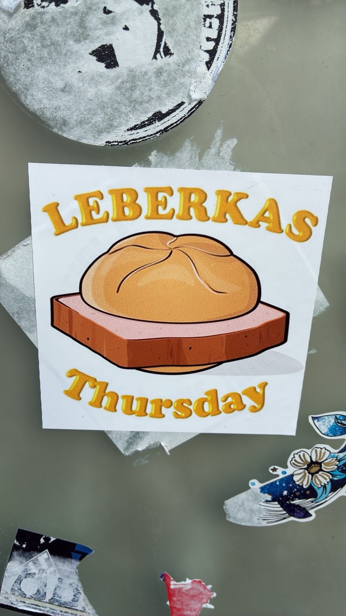 Leberkäse auf einem braunen rechteckigen Stück, belegt mit einem hellbraunen, runden Brötchen. Darüber steht in gelber Schrift „Leberkas“, darunter ebenfalls in Gelb „Thursday“. Der Hintergrund ist grau mit weiteren Aufklebern und Abnutzungsspuren.