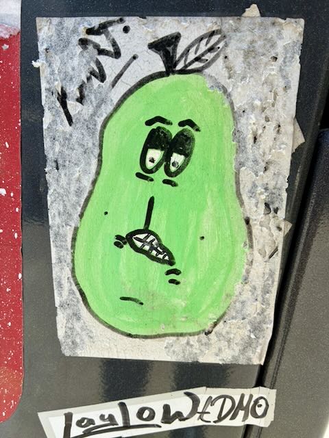 Handgezeichneter Sticker einer grünen Birne mit einfachen Comic-Gesichtszügen: große Augen, schmaler Nasenstrich, schiefes Gebiss. Oben ein kurzer Stiel mit Blatt. Hintergrund grau und abgenutzt, Kanten des Stickers teils aufgefasert; schwarze Kritzelei links oben.