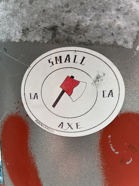 Runder Sticker mit rotem Beil in der Mitte, darum die Worte „SMALL AXE LA LA LA“ in schwarzer Schrift. Der Hintergrund ist weiß mit einigen Gebrauchsspuren.