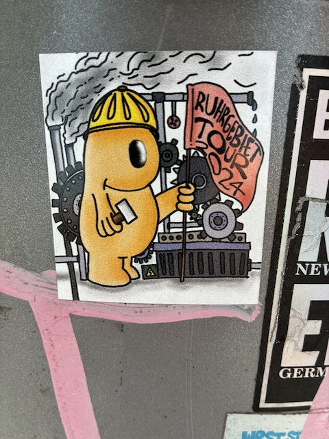 Knuffige gelbe Comicfigur mit Schutzhelm in industrieller Umgebung aus Zahnrädern und Schornsteinen. Figur hält ein Schild mit der Aufschrift „RUHRGEBIET TOUR 2024“ hoch.