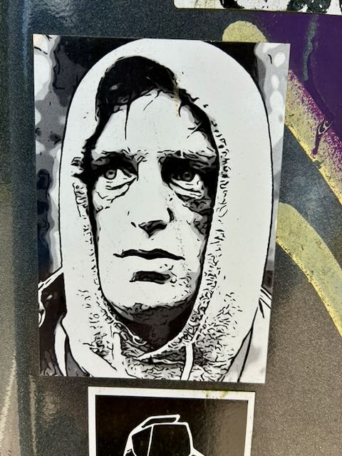 Schwarz-weißes Stencil-Poster einer Person mit Kapuze, frontal gezeigt. Markante Schatten um Augen und Wangen, neutraler Gesichtsausdruck. Der Druck ist rechteckig, leicht schief angebracht, auf einer Metallfläche mit farbigen Graffitistreifen im Hintergrund.
