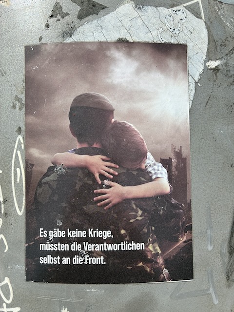 Ein Soldat in Tarnuniform umarmt einen Jungen vor zerstörter Stadtkulisse. Beide sind dem Betrachter den Rücken zugekehrt. Text unten: „Es gäbe keine Kriege, müssten die Verantwortlichen selbst an die Front.“