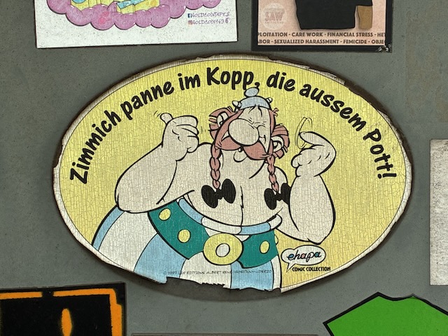 Comicfigur Obelix aus Asterix-Reihe mit blonden Zöpfen und blauem Helm zeigt Daumen nach oben. Comic-Schild trägt den Satz: „Zimnich panne im Kopp, die aussem Pott!“ und das Logo „Chap Comic Collection“.