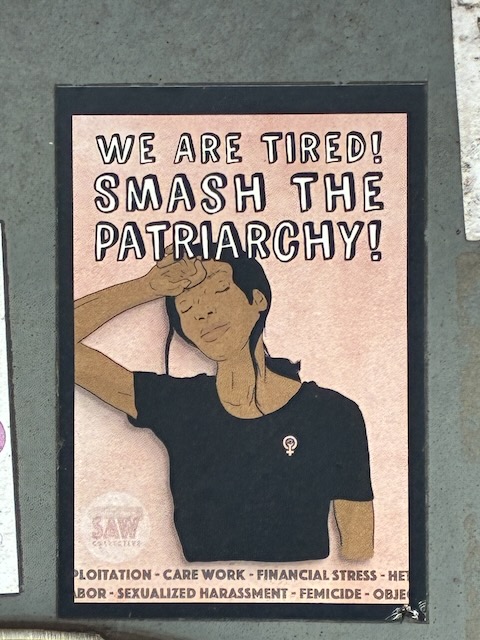 Frau mit langen schwarzen Haaren trägt ein schwarzes Shirt mit Feminsimus-Symbol und legt sich die Hand müde an die Stirn. Textbotschaft: "WE ARE TIRED! SMASH THE PATRIARCHY!". Weitere Worte am Rand thematisieren gesellschaftliche Missstände.