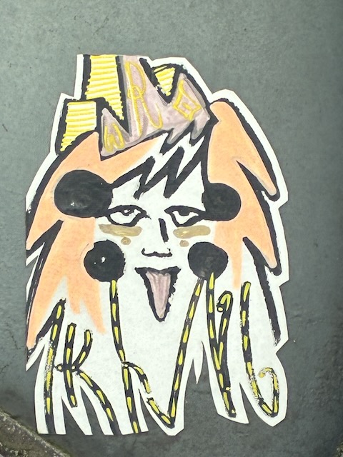 Gezeichnetes Gesicht mit orangefarbenen, unordentlichen Haaren, heraushängender Zunge und großen schwarzen Punkten als Wangen. Gelbe, gezackte Krone mit Aufschrift „KRAWALL“ auf dem Kopf; schwarze und gelbe Linien ziehen vom Gesicht nach unten.