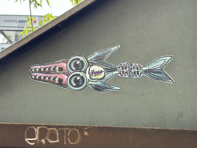 Gezeichneter, bunter Fisch mit metallisch schimmernden Farben, Flossen und großen Augen, die wie Schrauben wirken. Im Inneren des Körpers steht das Wort „Pirana“. Das Sticker befindet sich auf einer metallischen Oberfläche.