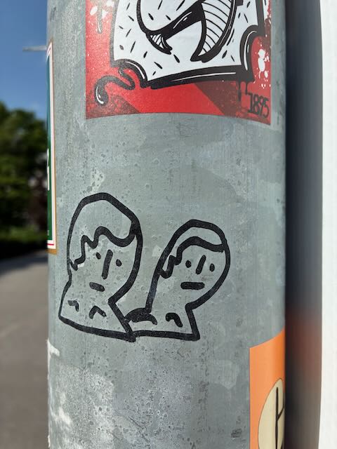 Eine einfache schwarze Strichzeichnung auf hellgrauem Untergrund zeigt zwei stilisierte Köpfe nebeneinander. Beide Figuren haben minimalistische Gesichter mit Punktaugen und neutralem Ausdruck.