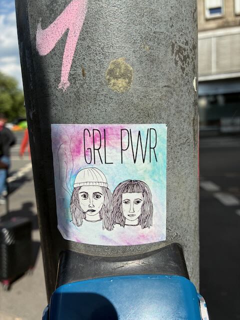 Sticker mit der Aufschrift „GRL PWR“ über zwei gezeichneten Frauenköpfen, die nebeneinander stehen. Der Hintergrund ist pastellfarben in Blau, Rosa und Violett gehalten.