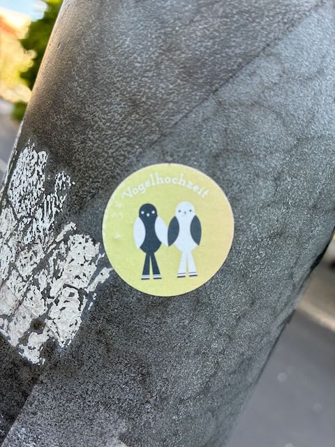 Runder Aufkleber auf gelbem Hintergrund mit zwei stilisierten Vögeln, einer schwarz, einer weiß, die nebeneinander stehen. Darauf steht „Vogelhochzeit“ in weißer Schrift.