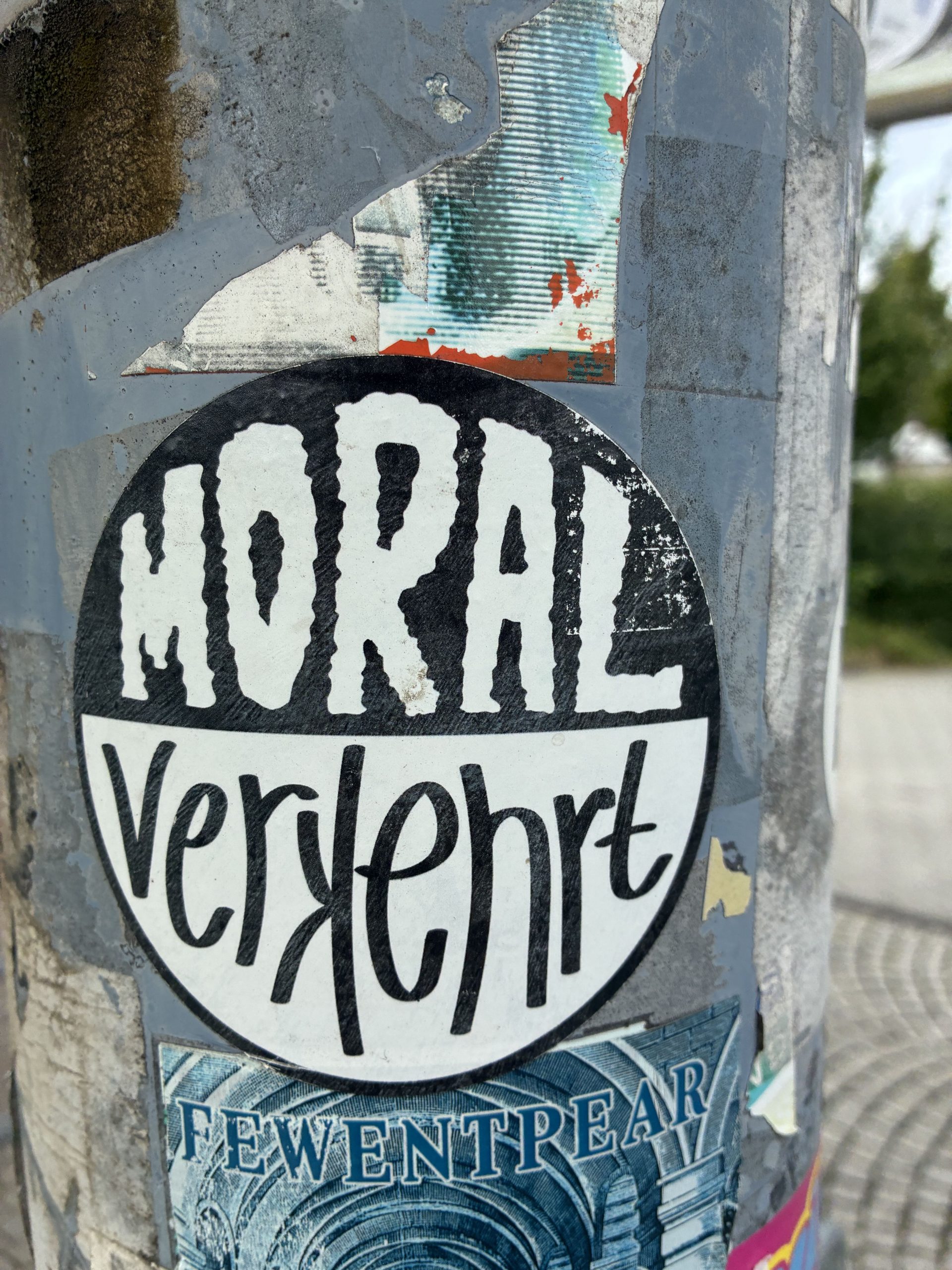 Runder schwarz-weißer Sticker auf einem grauen Laternenmast. Oben steht in weißen, ungleichmäßigen Großbuchstaben auf schwarzem Hintergrund „MORAL“, darunter in geschwungener schwarzer Schrift auf weißem Grund „verkehrt“. Der Aufkleber ist von anderen Stickern und abgerissenen Papierresten umgeben, die Oberfläche wirkt zerkratzt und verwittert.