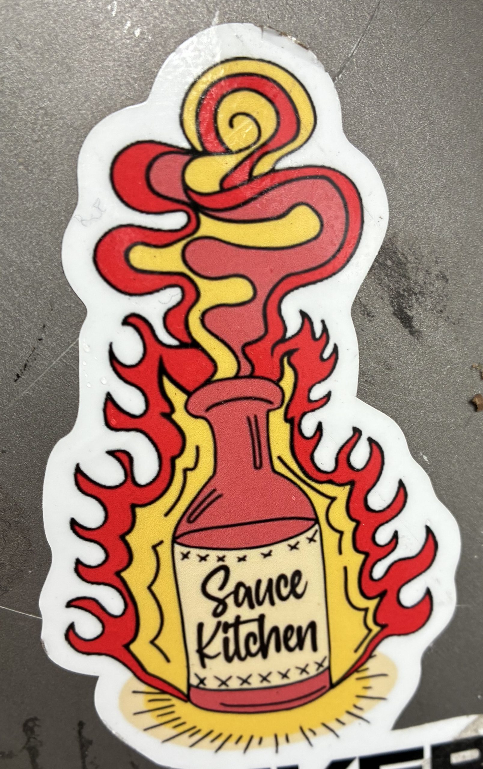 Konturgeschnittener Sticker mit einer roten Flasche, umgeben von stilisierten roten und gelben Flammen. Aus dem Flaschenhals steigt geschwungener Rauch in Rot und Gelb auf. Auf dem Etikett steht in schwarzer Handschrift „Sauce Kitchen“. Kräftige, kontrastreiche Farben auf weißem Hintergrund.
