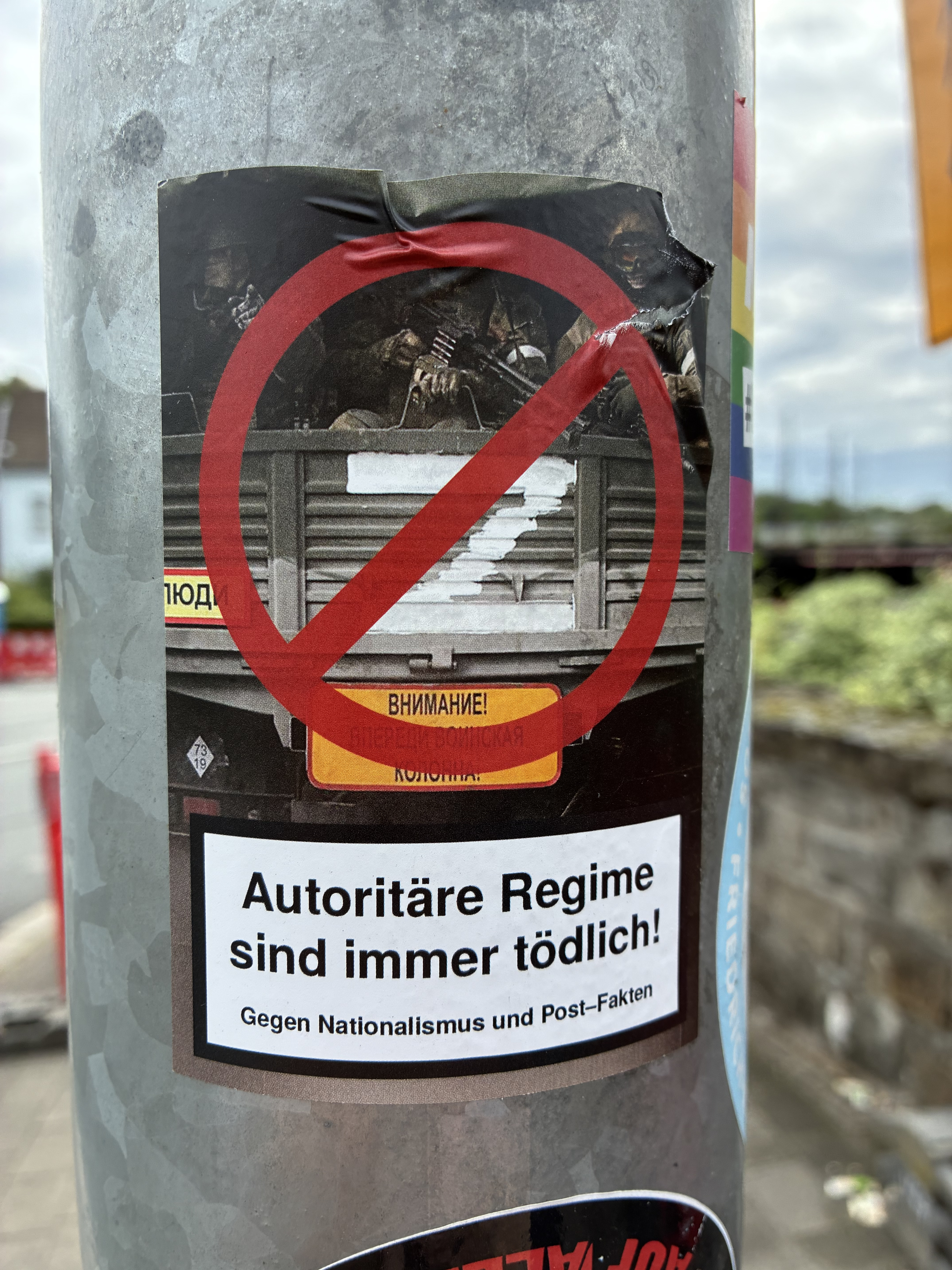 Bildbeschreibung: Auf einem grauen Metallpfosten klebt ein rechteckiger Sticker. Oben ist ein dunkles Bild mit Militärfahrzeug zu sehen, darüber ein rotes Verbotsschild in Form eines durchgestrichenen Kreises. Darunter steht in schwarzer Schrift auf weißem Hintergrund: „Autoritäre Regime sind immer tödlich!“ und kleiner: „Gegen Nationalismus und Post-Fakten“. Der Sticker ist leicht beschädigt und am Rand eingerissen.