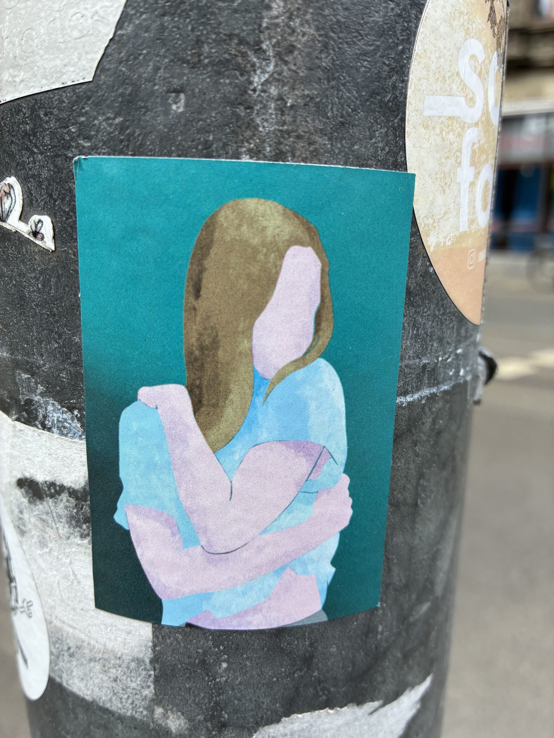 Rechteckiger Sticker auf einem dunklen Mast. Zu sehen ist eine stilisierte Person mit schulterlangem blondem Haar und blauem Oberteil vor türkisfarbenem Hintergrund. Das Gesicht ist ohne Details dargestellt. Die Figur umarmt sich selbst, die Arme in zarten Rosa- und Lilatönen gehalten.