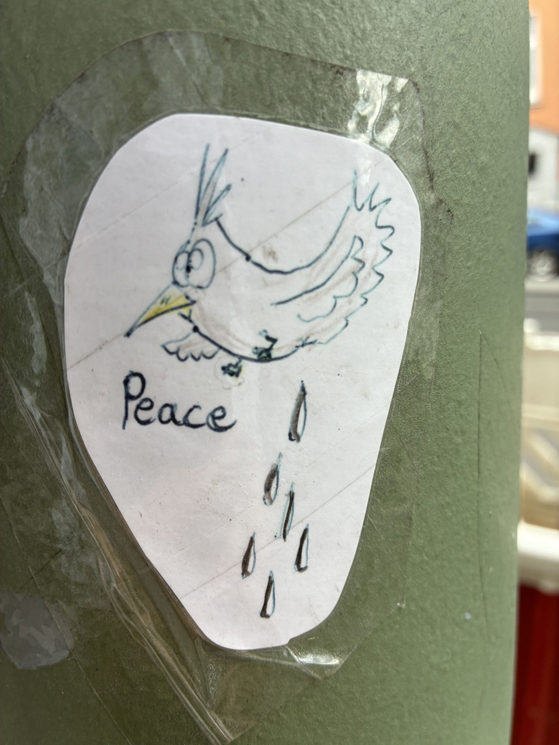 Unregelmäßig ausgeschnittener Sticker, mit transparentem Klebeband fixiert. Darauf eine einfache Zeichnung eines fliegenden Vogels mit gelbem Schnabel. Neben ihm steht das Wort „Peace“. Unter dem Vogel fallen mehrere tropfenförmige Zeichen nach unten. Die Linien sind locker und mit Stift in Blau und Schwarz gezeichnet.