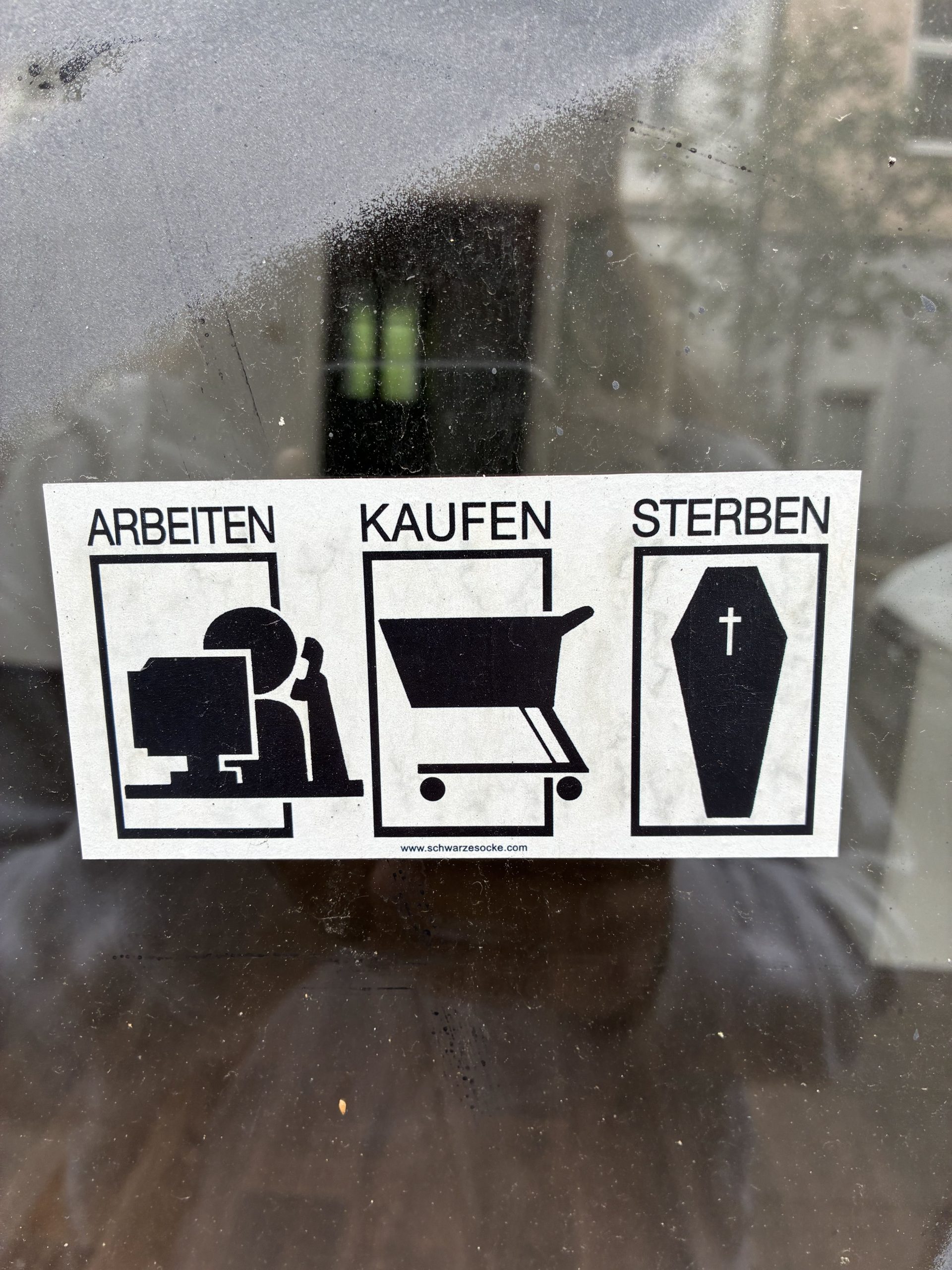 Rechteckiger schwarz-weißer Sticker auf Glas. Drei nebeneinander stehende Felder mit den Überschriften „ARBEITEN“, „KAUFEN“ und „STERBEN“. Darunter einfache Piktogramme: eine Person am Computer, ein Einkaufswagen und ein Sarg mit Kreuz. Die Darstellung wirkt wie eine Abfolge des Lebens in klaren Symbolen.