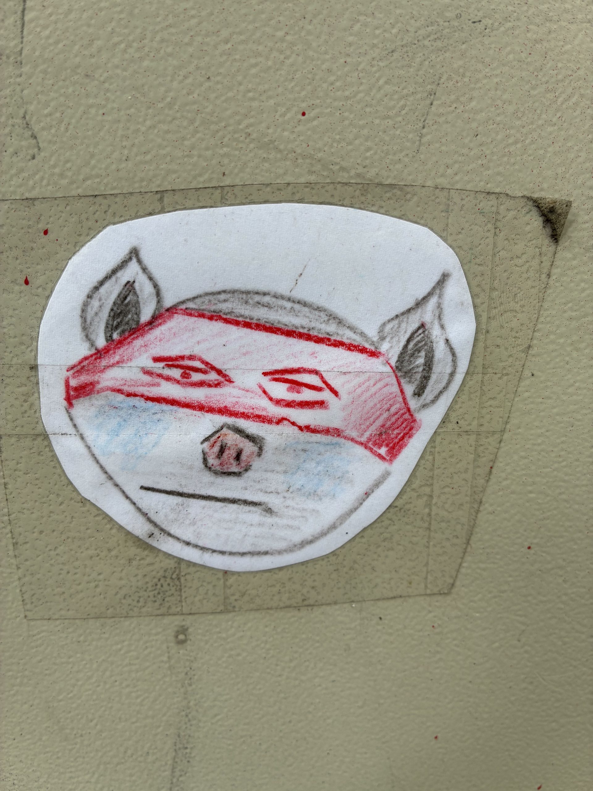 Runder, mit Klebeband befestigter Sticker auf hellgrünem Untergrund. Zu sehen ist ein handgezeichnetes Tiergesicht mit spitzen Ohren und kleiner Schnauze. Über den Augen trägt es eine rote, maskenartige Fläche. Die Zeichnung wirkt skizzenhaft, mit Buntstift in Grau, Rot und etwas Blau schattiert.