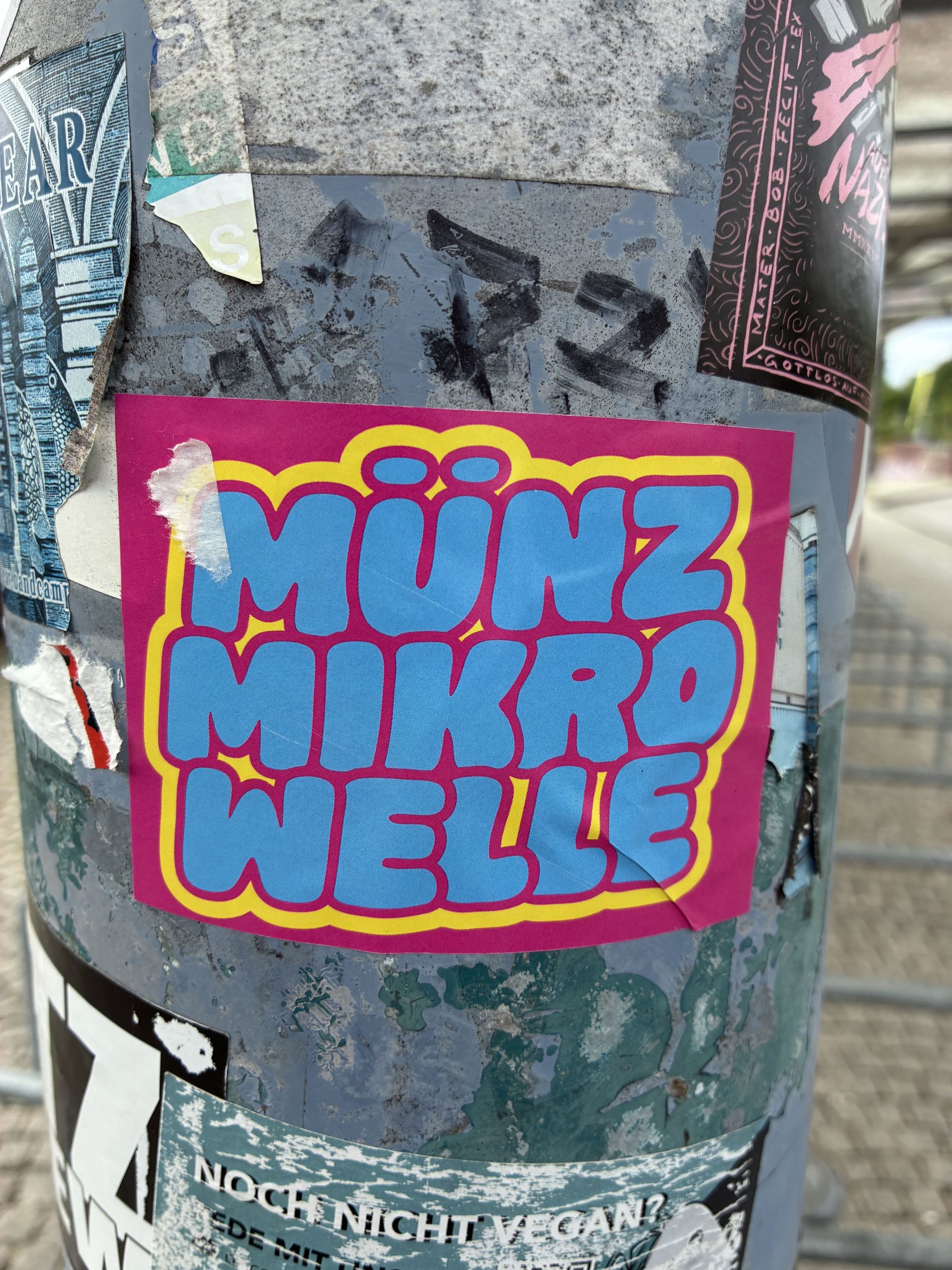 Ein knallpinkfarbener Sticker mit blauem, comicartigem Schriftzug "MÜNZ MIKRO WELLE" in Großbuchstaben, umrandet von einem gelben Streifen. Der Sticker klebt auf einem metallenen Pfosten, umgeben von anderen überklebten Aufklebern.