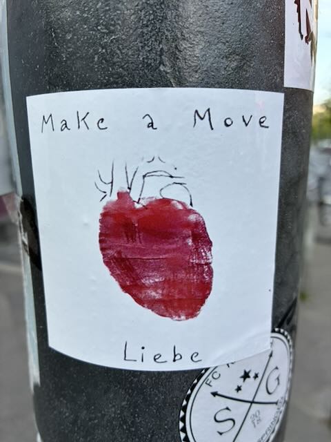 Foto eines rechteckigen weißen Stickers auf einer dunklen Metallfläche. In der Mitte ist ein stilisiertes rotes anatomisches Herz, teils gemalt, teils skizziert. Oberhalb steht in handschriftähnlicher schwarzer Schrift „Make a Move“, darunter mittig das Wort „Liebe“. Rechts und links sind Teile weiterer überklebter Aufkleber zu sehen.