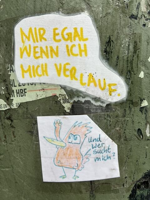 Auf einer abgenutzten, graugrünen Fläche kleben zwei Sticker. Oben ein weißer, unregelmäßig geformter Aufkleber mit gelber Handschrift „MIR EGAL WENN ICH MICH VERLAUF.“. Darunter ein kleiner rechteckiger Zettel mit einer einfachen, bunten Kritzel-Figur mit Schnabel und erhobener Hand; daneben in Blau der handschriftliche Text „und wer sucht mich?“.