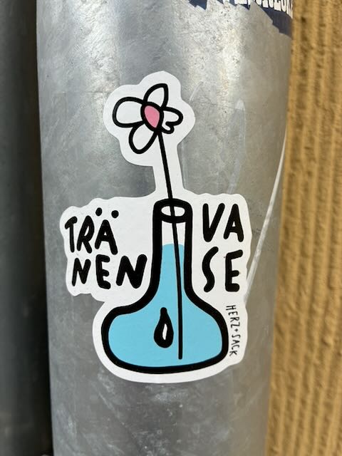 Auf einer grauen Metallfläche klebt ein weiß umrandeter Sticker. Darauf ist ein stilisiertes, blau gefülltes Gefäß mit schwarzem Rand und einem einzelnen Tropfen im Inneren zu sehen. Aus der Öffnung ragt ein dünner Stiel mit einfacher Blüte, weiß mit rosa Mitte. Um das Gefäß steht in schwarzer Handschrift das Wort „TRÄNENVASE“, am Rand klein „HERZ.SACK“.