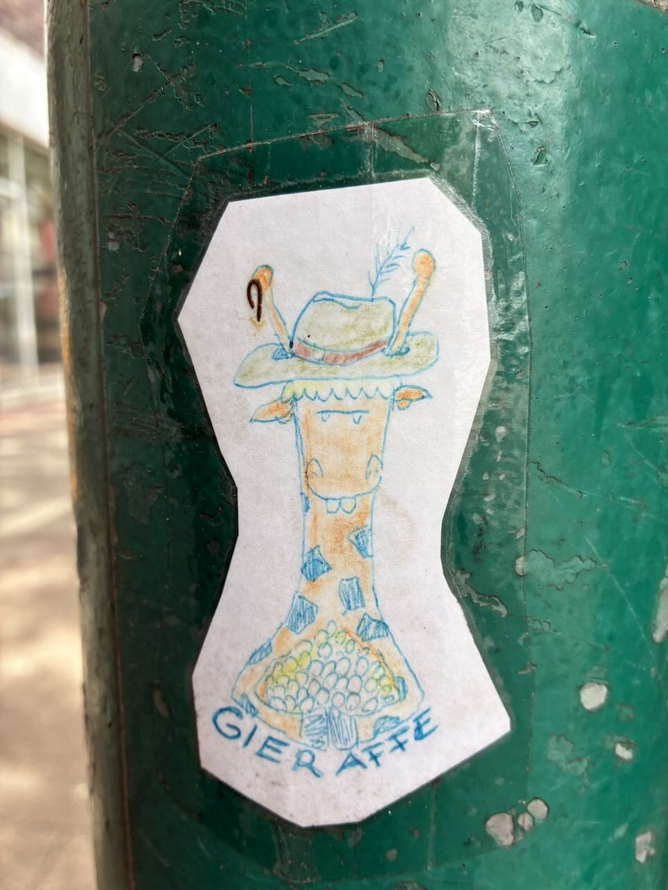 Handgezeichneter Sticker an einer grünen Säule. Zeigt eine Giraffe mit einem Hut, auf dem ein Vogel sitzt. Unter der Giraffe steht in blauer Schrift "GIER AFFE". Der Sticker ist leicht beschädigt und hat abblätternde Ränder.