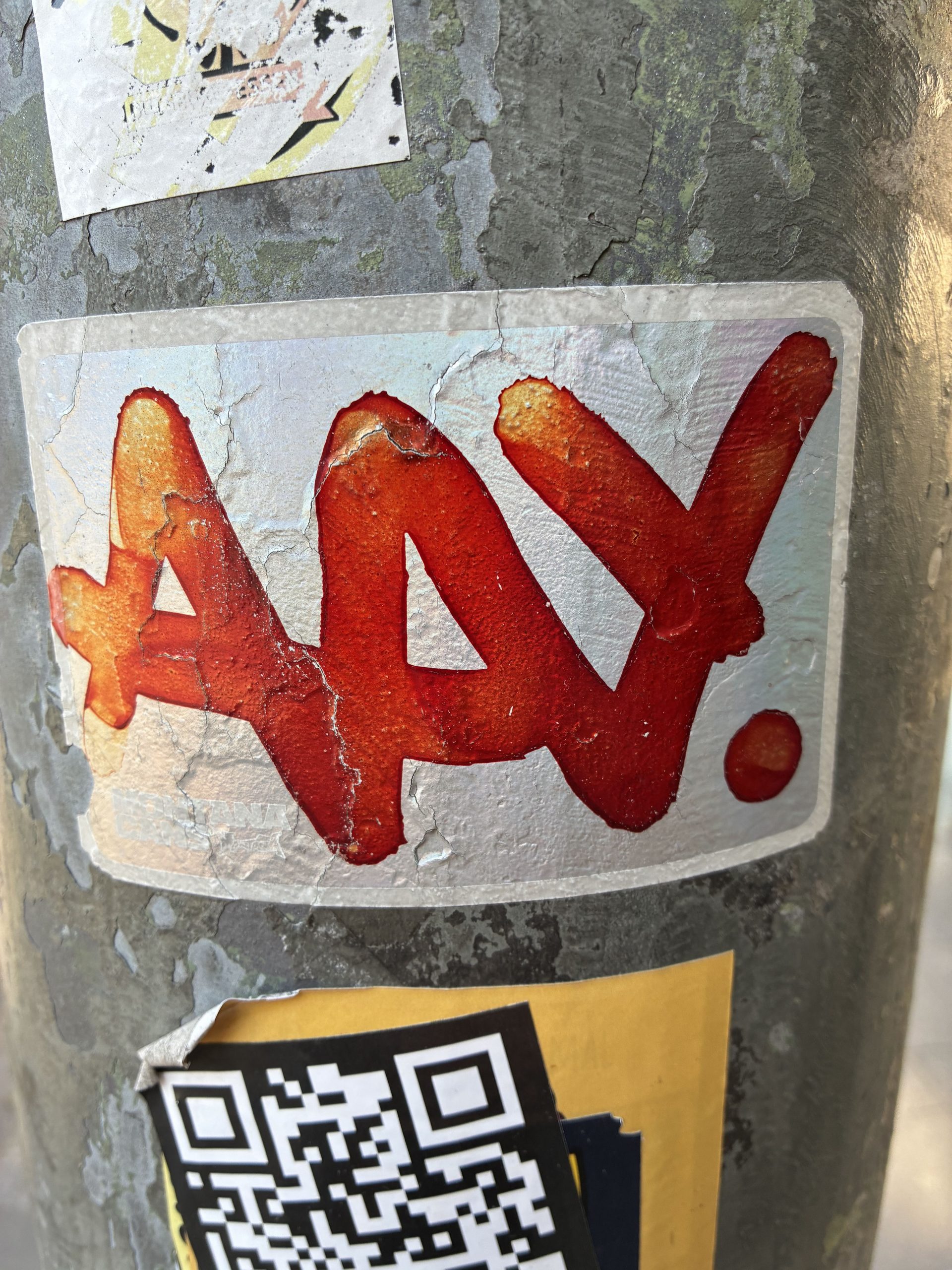 Rechteckiger Sticker mit weißem Hintergrund und grob aufgetragener, glänzend roter Farbe. Darauf steht in großen, verlaufenden Buchstaben „AAAY.“ mit einem Punkt am Ende. Die Ränder sind rissig und abgeblättert. Der Sticker klebt auf einem grauen, verwitterten Laternenmast; darunter ist teilweise ein QR-Code-Aufkleber zu sehen.
