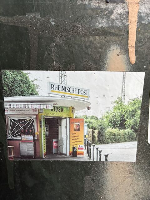 Foto-Sticker eines kleinen Kiosks an einer Straße. Über dem flachen Dach hängt ein Schild „RHEINISCHE POST“. Unter der Markise steht „Kiosk“ und weitere Schrift. Davor ein gelbes Werbeschild mit Text, Getränkekisten und ein Mülleimer. Rechts führt der Blick auf eine leere Straße mit grünen Büschen und Bäumen, oben sind Masten oder Stromleitungen zu sehen.