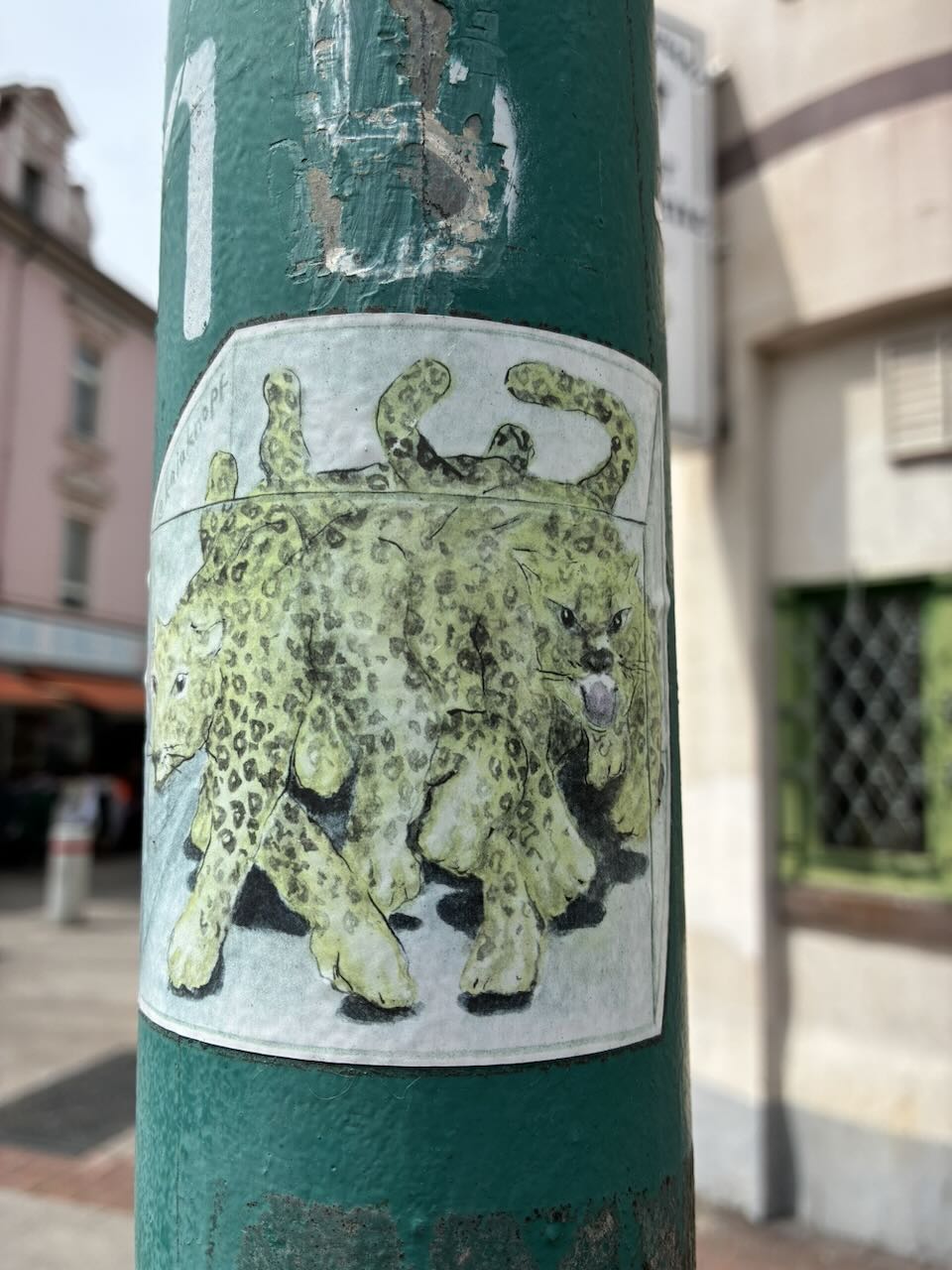 Sticker auf einer grünen Säule in urbaner Umgebung. Der Sticker zeigt zwei gelbe Leoparden mit schwarzen Flecken, die nebeneinander laufen. Der Hintergrund des Stickers ist weiß, und der Sticker ist leicht beschädigt, mit sichtbaren Rissen und abblätternden Rändern.