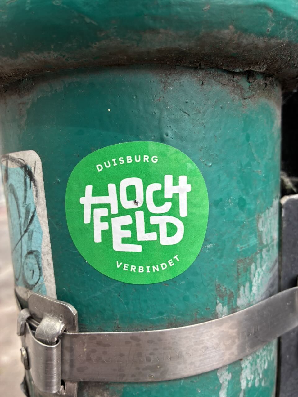 Runder, grüner Sticker auf einem abgenutzten, türkisfarbenen Metallbehälter. Der Sticker zeigt weiße Schrift: "DUISBURG HOCHFELD VERBINDET". Der Behälter ist mit einem Metallband gesichert.