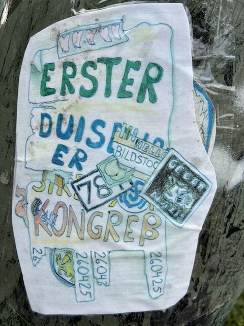 Auf einer grauen Oberfläche klebt ein weißer, unregelmäßig ausgeschnittener Sticker mit handgezeichneten, farbigen Buchstaben und kleinen Bildern. Groß steht „ERSTER DUISER KONGREB“, dazu mehrere kleine Kästchen mit Zahlen wie „78“ und Datumsangaben „26.04.25“. Überlagert sind mehrere kleine, rechteckige Aufkleber mit weiteren Zeichnungen und Schriftzügen.