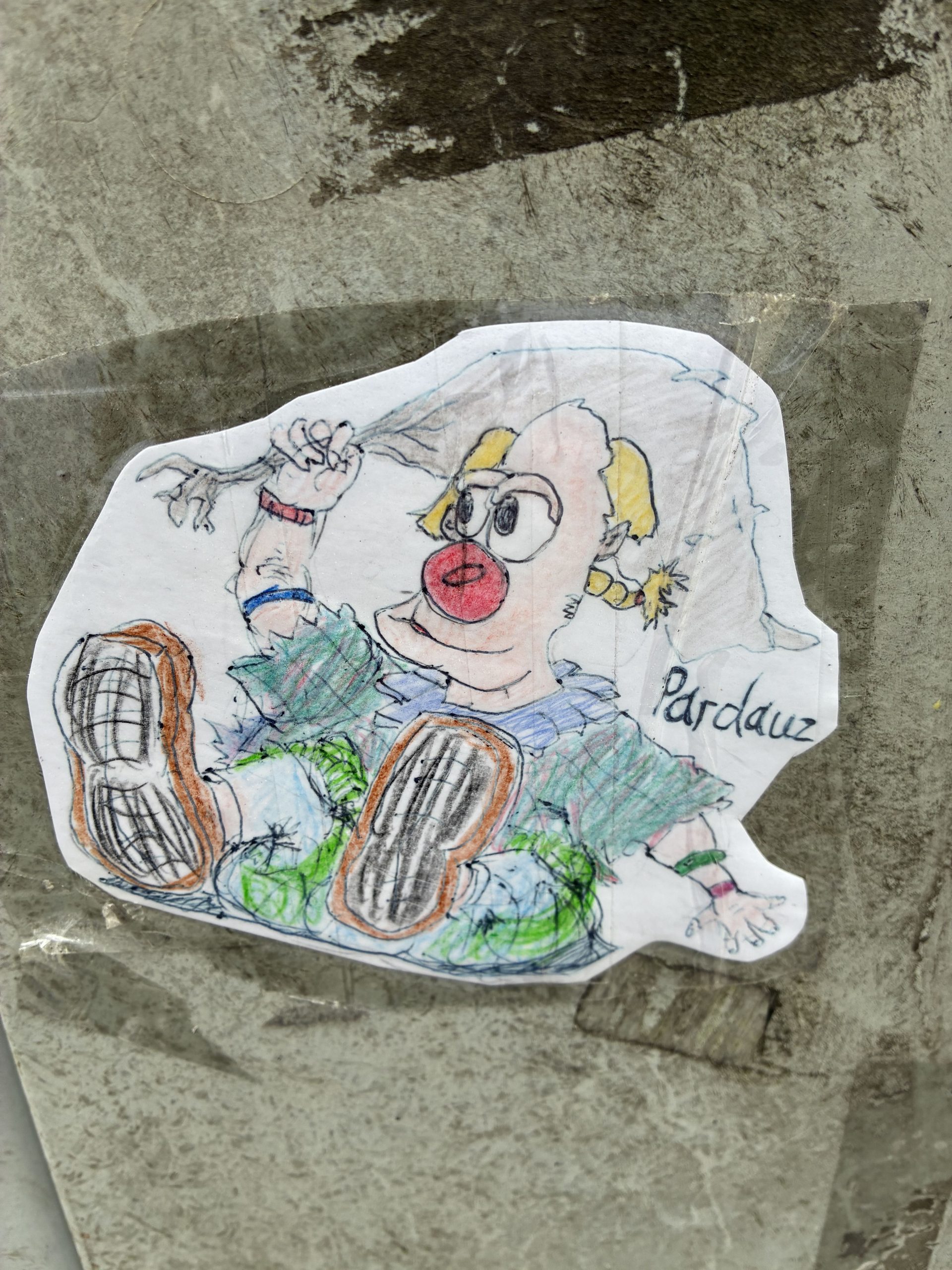 Ein ausgeschnittener, handgezeichneter Sticker zeigt einen Clown mit roter Nase, gelben Haaren und Zipfelmütze. Er liegt auf dem Rücken, die großen Schuhsohlen mit Profil nach vorn gestreckt. Die Kleidung ist grün und blau schraffiert. Der Sticker ist mit transparentem Klebeband auf einen grauen, verschmutzten Mast geklebt. Daneben steht handschriftlich ein Name.