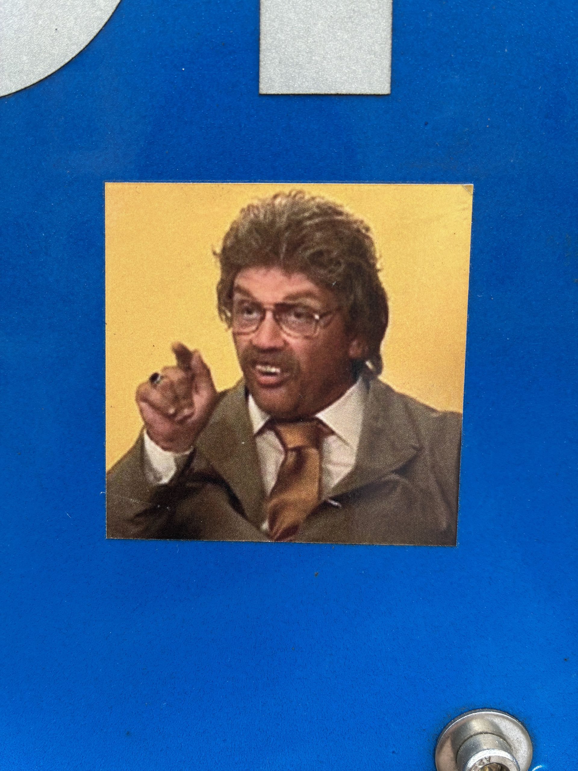 Quadratischer Sticker auf blauem Untergrund zeigt einen Mann mit graubrauner Frisur, Brille und Schnurrbart („Horst Schlämmer“). Er trägt Anzug und Krawatte und zeigt mit ausgestrecktem Finger direkt nach vorn. Sein Mund ist geöffnet, als würde er sprechen oder schimpfen. Unten rechts ist ein rundes Metallelement des Untergrunds sichtbar.