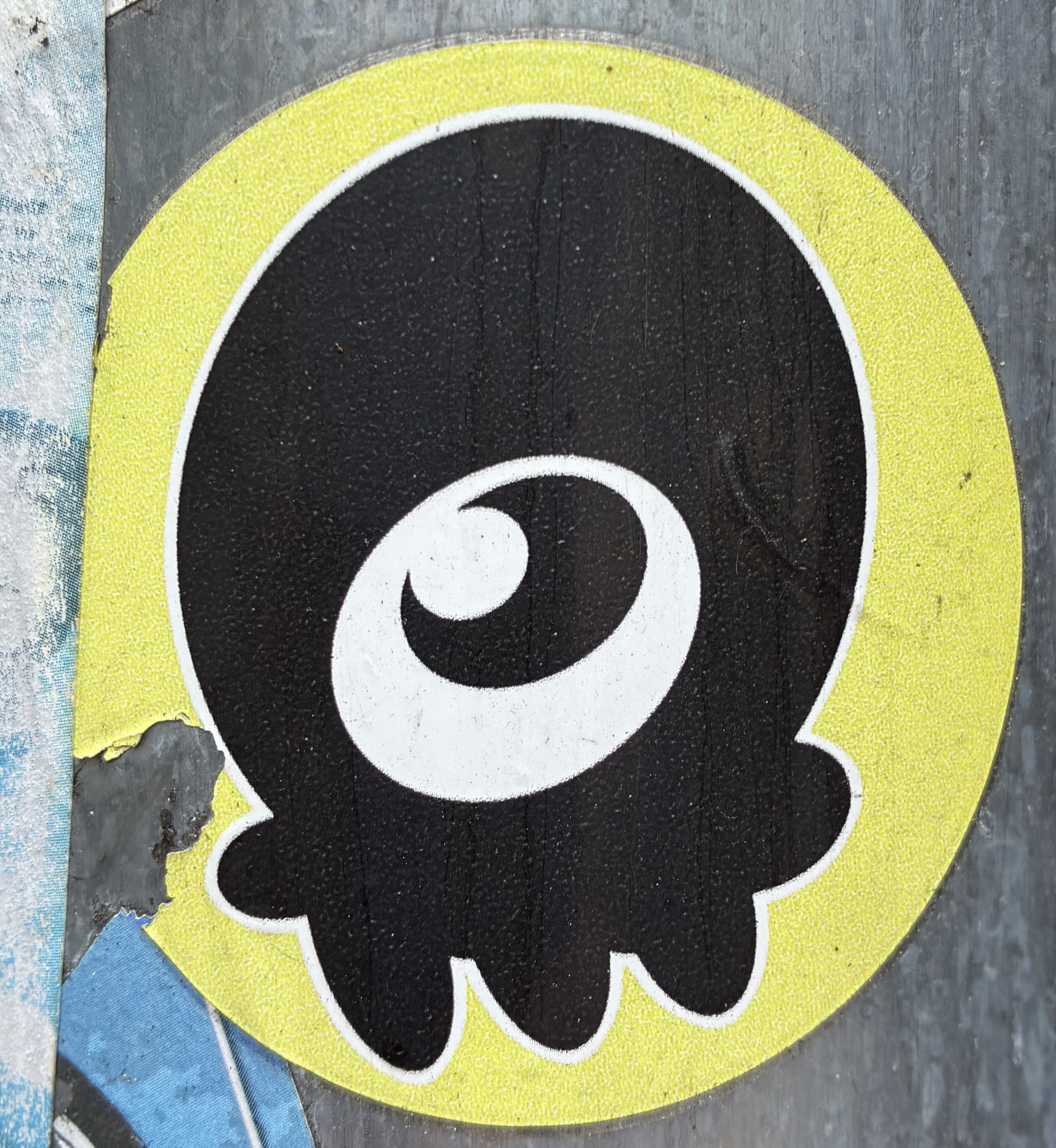 Runder Sticker mit grellgelbem Rand. In der Mitte auf schwarzem Grund eine vereinfachte, tropfenförmige Figur mit einem großen weißen Auge, das nach oben blickt. Unten endet die Form in mehreren kurzen, fransenartigen Ausläufern. Der Aufkleber klebt auf einem grauen Mast und ist am Rand leicht beschädigt.