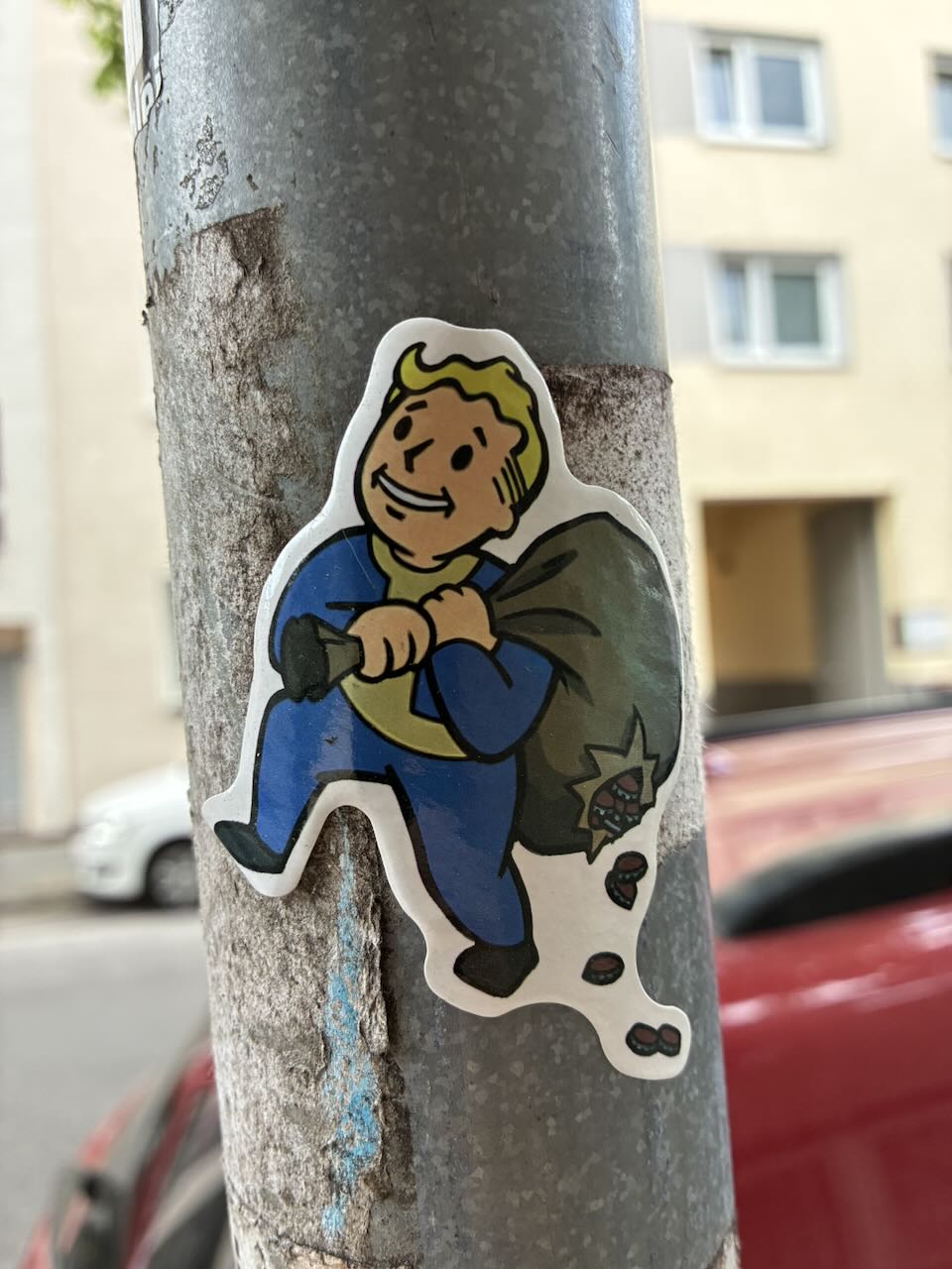 Sticker an einer grauen Säule in urbaner Umgebung. Zeigt eine lächelnde Comic-Figur mit gelber Haut, blonden Haaren und blauem Overall, die einen großen Beutel trägt. Die Figur hat eine freudige Mimik. Im Hintergrund sind Gebäude und ein geparktes Auto zu sehen.