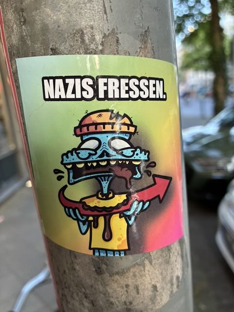 Rechteckiger bunter Sticker auf einem Laternenpfahl. Oben steht in weißer Blockschrift mit schwarzer Umrandung „NAZIS FRESSEN.“. Darunter eine Comicfigur mit blauem Kopf und Krone, die ein rotes Pfeilsymbol wie eine Wurst im Mund hat. Die Figur hält einen gelben Teller oder eine Scheibe, aus der gelbe Masse tropft. Hintergrund ist ein Farbverlauf in Gelb, Grün und Pink.