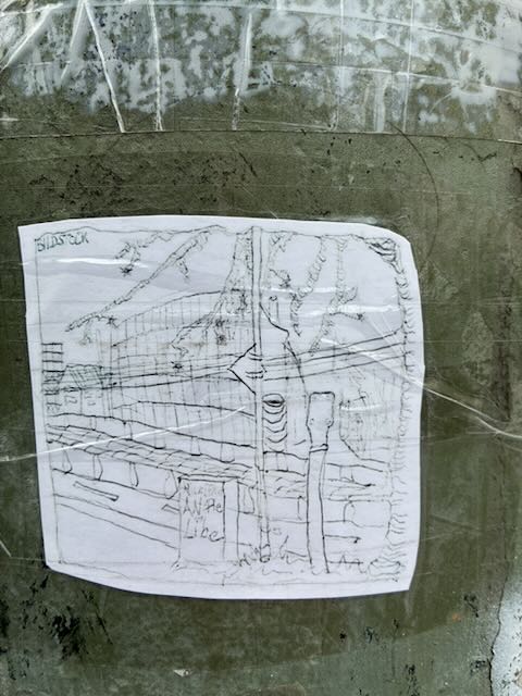 Auf einer grauen, leicht ramponierten Oberfläche klebt ein weißes Blatt Papier, mit durchsichtigem Klebeband befestigt. Darauf ist eine feine Bleistiftskizze zu sehen: Straße mit Geländer im Vordergrund, Laternenmast und Verkehrsschild, dahinter ein längliches Gebäude mit vielen Fenstern sowie angedeutete Bäume und Berge.