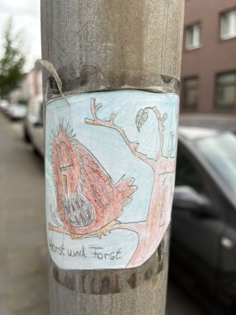 Foto eines an einem grauen Pfahl befestigten, handgezeichneten Aufklebers. Zu sehen ist ein rötlich brauner Vogel mit grauer Brust, der auf einem Ast eines kahlen Baumes sitzt. Hintergrund hellblau, rechts ein kleiner grüner Blätterzweig. Unten auf dem Aufkleber steht handschriftlich „Horst und Forst“. Im unscharfen Hintergrund geparkte Autos und Häuserfassaden an einer Straße.