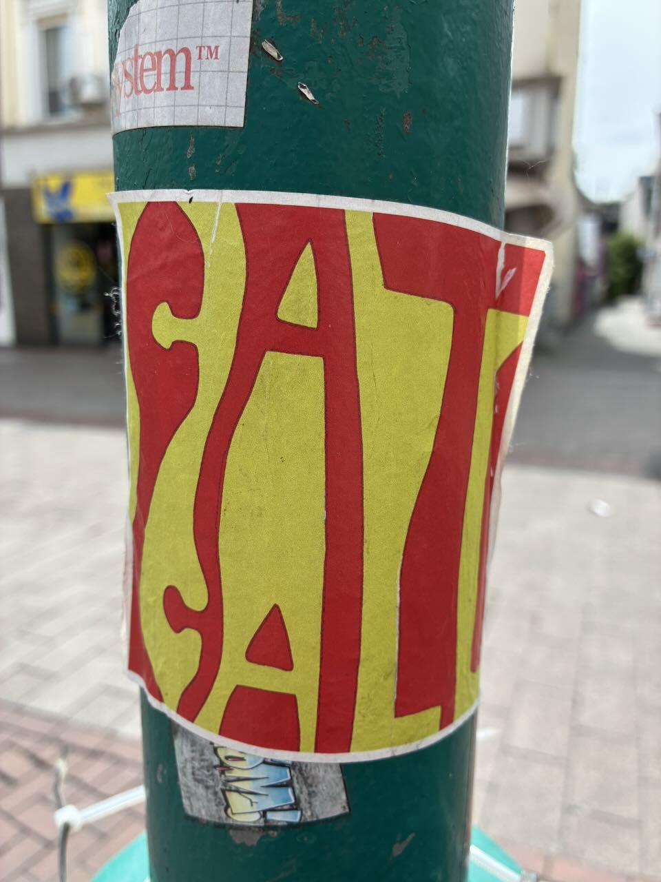 Sticker auf einer grünen Säule in einer Stadtumgebung. Der Sticker zeigt die gelb-roten Streifen der katalanischen Flagge und enthält die Aufschrift "CAT" in großen, roten Buchstaben auf gelbem Grund. Der Sticker ist teilweise beschädigt und hat abstehende Ränder. Im Hintergrund sind weitere Aufkleber und ein "System"-Logo zu sehen.