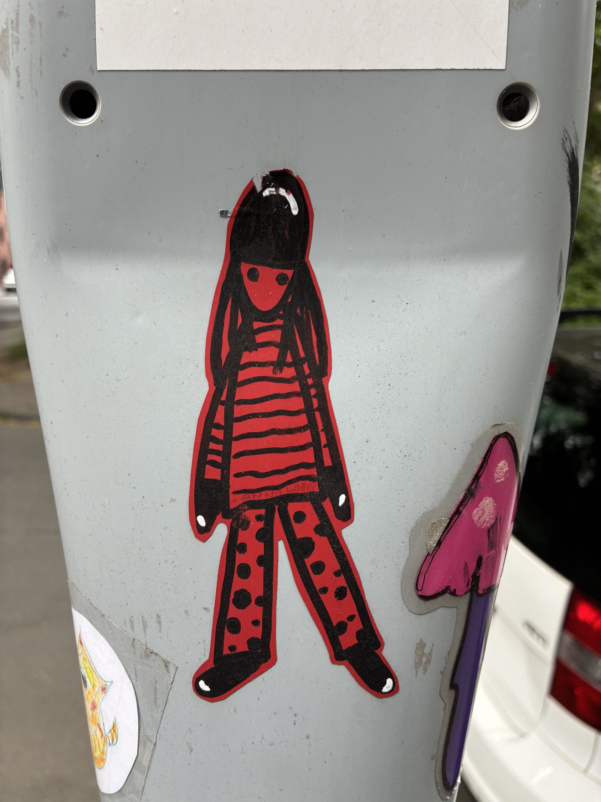 Schmaler Sticker auf einem grauen Mast zeigt eine stilisierte, rot-schwarze Figur mit langen Haaren. Sie trägt ein rot gestreiftes Oberteil, weite Hosen mit Punkten und schwarze Schuhe. Gesicht und Details sind stark vereinfacht, die Konturen dick schwarz umrandet. Die Haltung ist frontal und leicht breitbeinig.