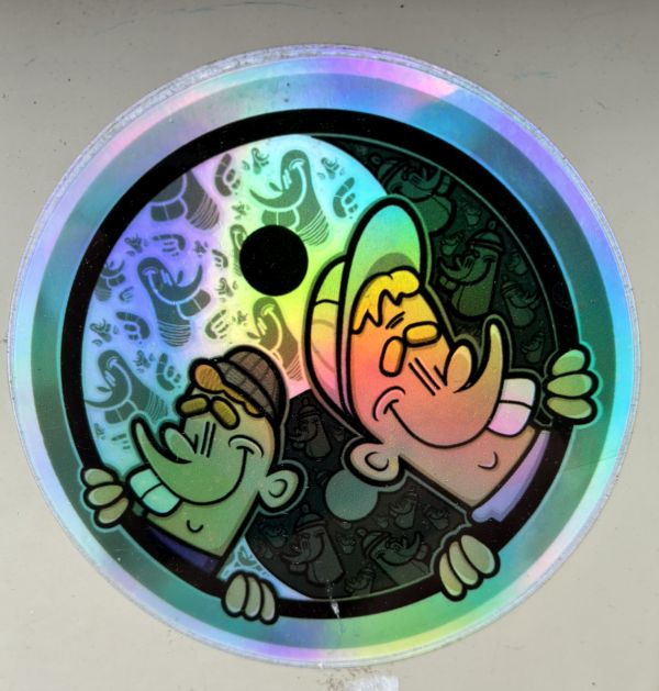 Runder Sticker mit holografischem Rand in Regenbogenfarben. Im Zentrum ein Yin-Yang-Symbol mit schwarzem Rand und schwarzem Punkt. In die zwei Hälften des Symbols sind je eine Cartoon-Figur integriert: links eine grüne Figur mit brauner Mütze und großer Nase, rechts eine Figur mit gelbem Haar und orangefarbenen Backen. Der Hintergrund beider Hälften zeigt ein Muster aus kleinen Cartoon-Motiven.